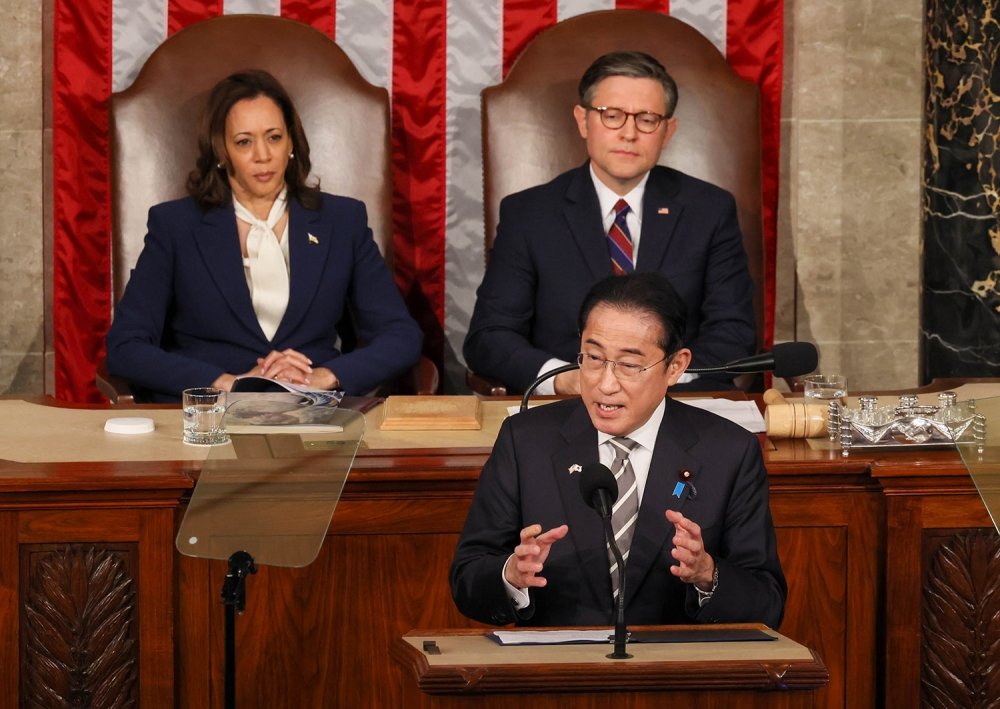 Prime Minister Kishida stands at a lecturn giving a speech, Vice President Harris and Speaker Johnson are seated behind him.